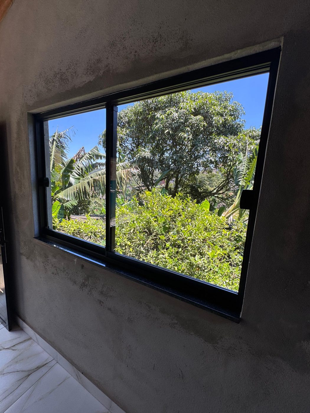 Janela de alumínio com perfil preto instalada em parede de concreto, com vista para a vegetação